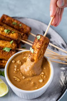 someone dipping some food into a bowl with skewers on the plate next to it