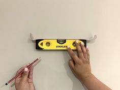 two hands holding pencils near a yellow and black ruler with holes in the wall