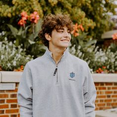 a young man standing in front of a brick wall with flowers and trees behind him