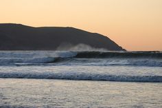 the sun is setting over an ocean with waves crashing in front of a rock formation