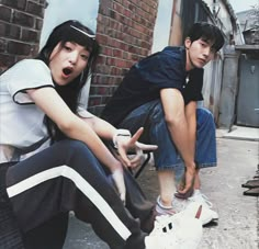 two young people sitting on the ground with their hands in their pockets and one is pointing at something