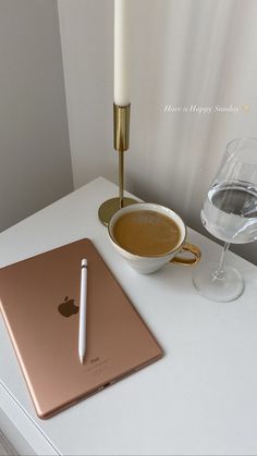 an apple laptop, pen and glass of wine sit on a table next to a candle