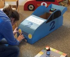 a woman kneeling down next to a child's car shaped cardboard box on the floor