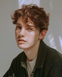 a young man with curly hair sitting in front of a white wall and looking off to the side