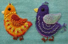 two embroidered birds sitting next to each other on a blue cloth covered surface, one is yellow and the other is purple