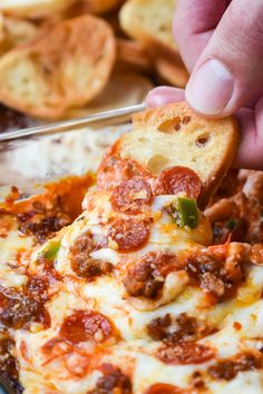 a hand holding a piece of bread with pepperoni and cheese on it, over a pan of baked crackers