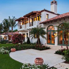 a large white house with lots of windows and plants in the front yard, surrounded by palm trees