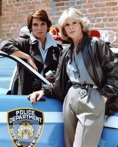 two women standing next to each other in front of a police car