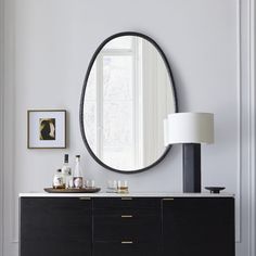 a large round mirror hangs on the wall above a black dresser and sideboard in a white room