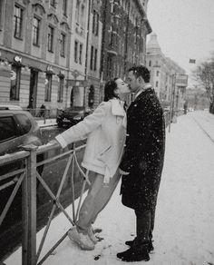 a man and woman kissing in the snow by a fence on a city street with buildings behind them