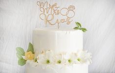 a wedding cake with white flowers and a gold monogram on the top is shown