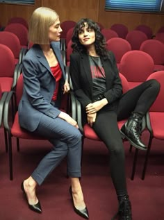 two women sitting on chairs in front of an empty auditorium with red seats behind them