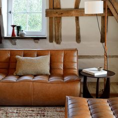 a brown leather couch sitting in front of a window next to a table with a lamp on it