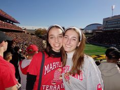 two girls in red and gray shirts at a football game, one is holding her arm around the other's shoulder