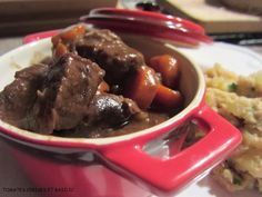 a red bowl filled with meat and vegetables on top of a white plate next to mashed potatoes