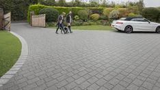 two people walking in front of a white car on a paved driveway next to bushes