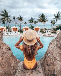 a woman in a yellow swimsuit and hat looking out over the pool at palm trees