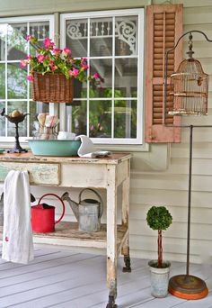 an old table with flowers and watering cans on it in front of a window filled with potted plants