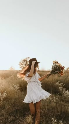 a woman in a white dress and cowboy hat walking through tall grass with flowers on her head