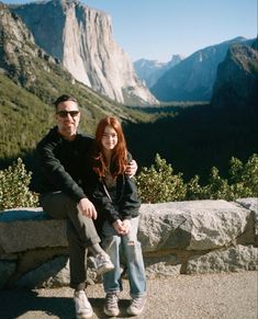 a man and woman sitting on top of a stone bench in front of some mountains