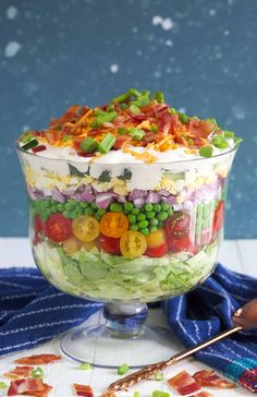 a layered salad in a glass bowl with bacon, peas and tomatoes on the side