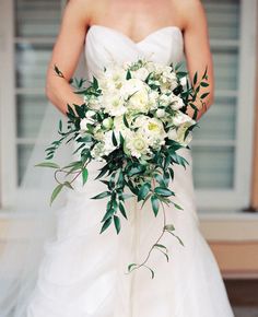 a bride holding a bouquet of white flowers and greenery on her wedding day in front of a window