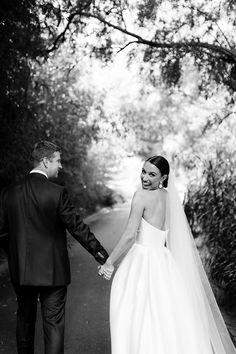 a bride and groom holding hands while walking down a path in front of some trees