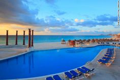 an empty swimming pool next to the beach with blue chairs and umbrellas around it