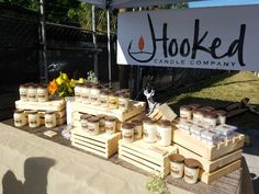 many jars of candles are on display at the outdoor market table with sign in background