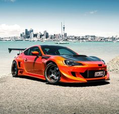 an orange sports car parked in front of the water with a cityscape in the background