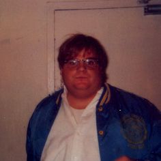 a man wearing glasses and a blue jacket standing in front of a door with his hand on his hip