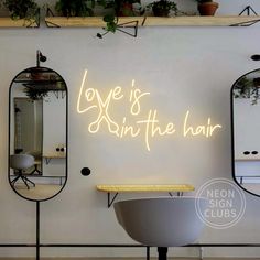 love is in the hair neon sign on a wall next to two mirrors with plants