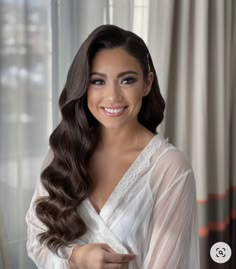 a woman with long dark hair wearing a white robe and smiling at the camera, in front of curtains