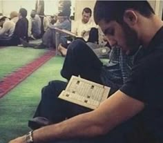 a man sitting on the floor reading a book with other people in the room behind him