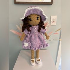 a crocheted doll in a purple dress and hat on a white table next to a painting