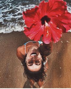 a woman holding a large red flower on top of her head in front of the ocean