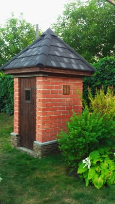 a small brick outhouse sitting in the middle of some grass and bushes with trees behind it