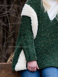 a woman wearing a green and white sweater standing next to a wooden fence with her hands in her pockets