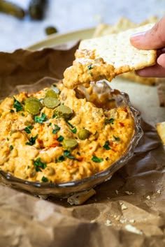 a hand holding a cracker over a bowl of hummus and pickle chips