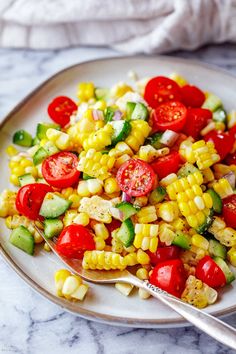 a white plate topped with corn, tomatoes and cucumbers next to a fork