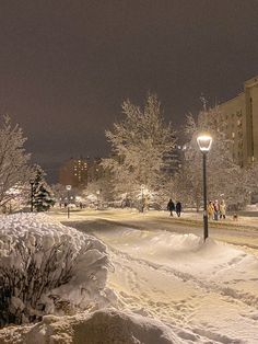 people walking down a snowy street at night with snow on the ground and buildings in the background