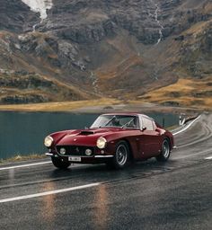 an old red sports car driving down the road in front of some mountains and water