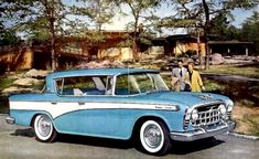 an old blue and white car parked in front of a house with two men standing next to it