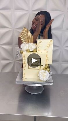 a woman sitting in front of a cake on top of a silver table next to a white wall