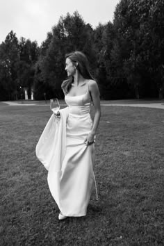 a woman in a long white dress holding a wine glass and walking through the grass