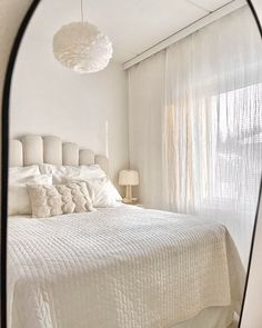 a large white bed sitting under a window next to a lamp and a mirror on the wall