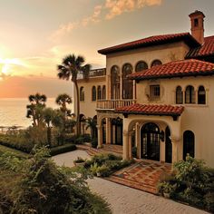 a large house sitting on top of a lush green hillside next to the ocean at sunset