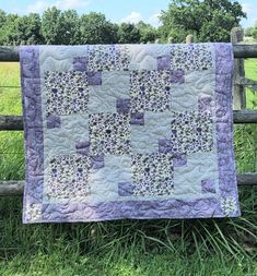 a purple and white quilt hanging on a wooden fence in front of a field with green grass