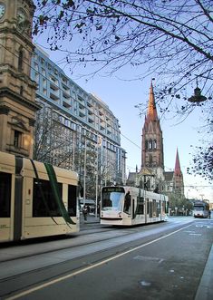 two white buses driving down a street next to tall buildings and a church steeple