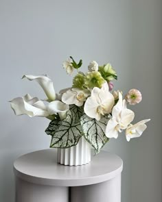 a white vase filled with lots of flowers on top of a table next to a gray wall